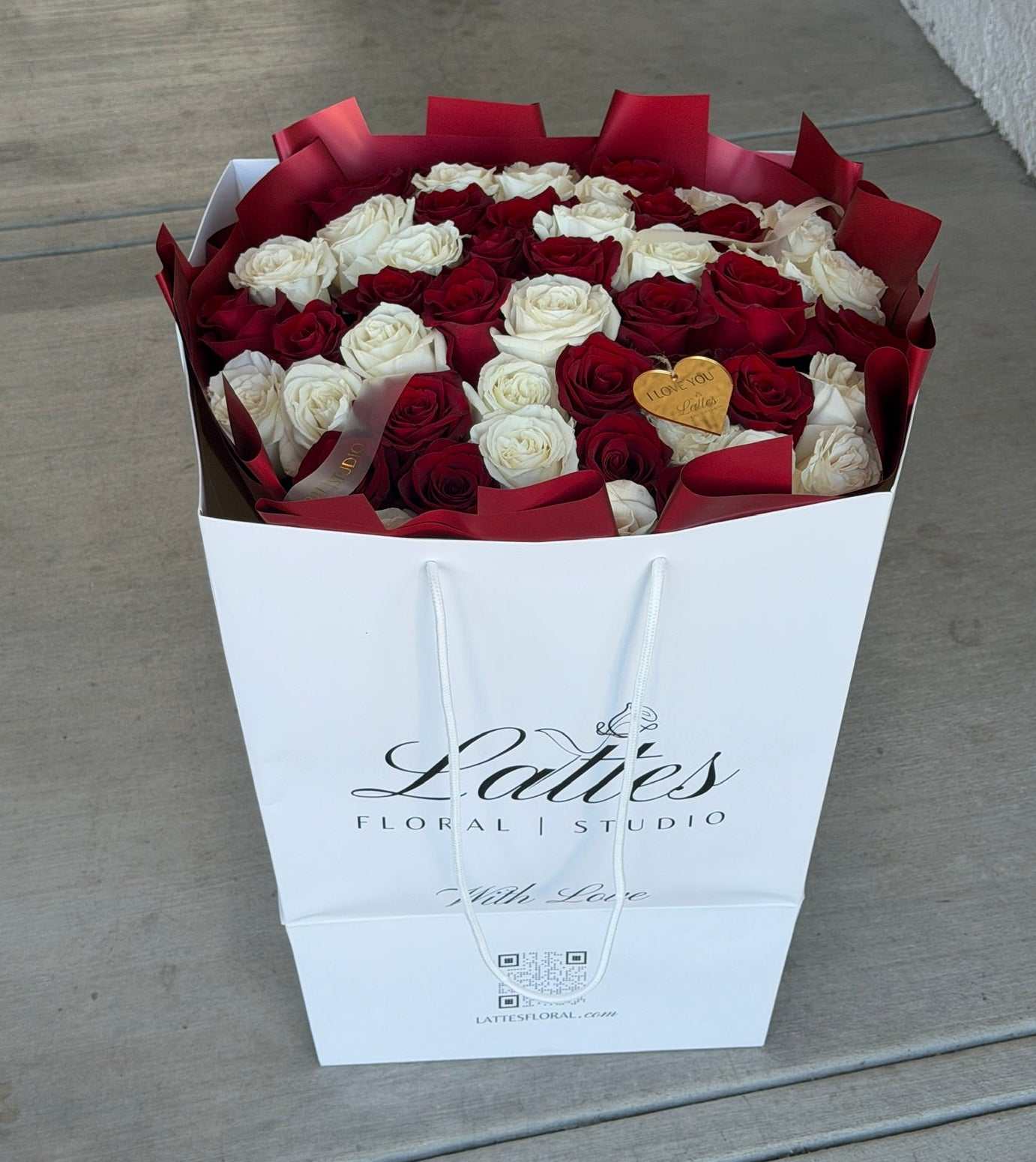 A large bouquet of red and white roses in a white gift box with red tissue paper, placed on a concrete floor. The box is branded 'Ladies Floral Studio'.