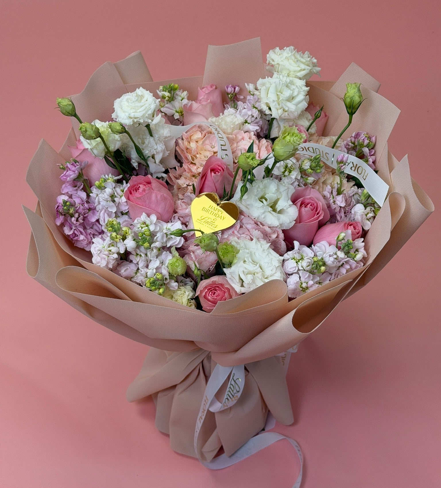 A hand-tied bouquet of pink and white flowers wrapped in beige paper, featuring roses, carnations, and lilac sprigs with a small gold heart-shaped tag, set against a solid pink background.