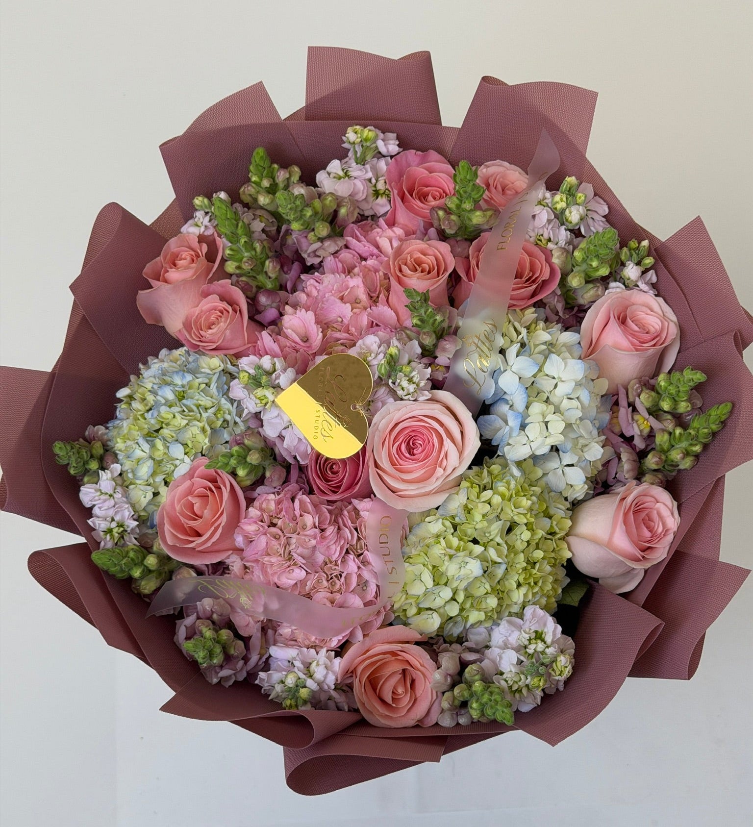 A top-down view of a floral bouquet wrapped in dark brown paper, featuring pink roses, light pink hydrangeas, pale blue hydrangeas, and green filler flowers, with a small gold heart-shaped card in the center.