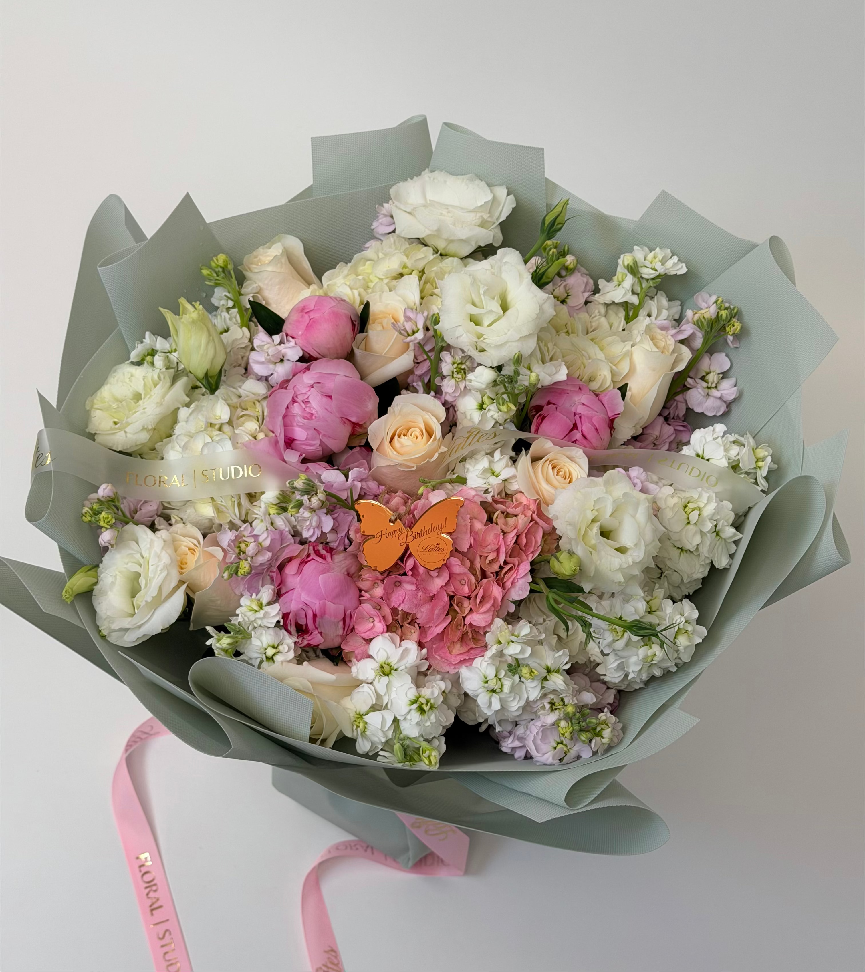 A beautifully arranged floral bouquet featuring white and pink flowers, wrapped in a sage green paper wrapper with a pink ribbon that reads 'Floral Delivery'.