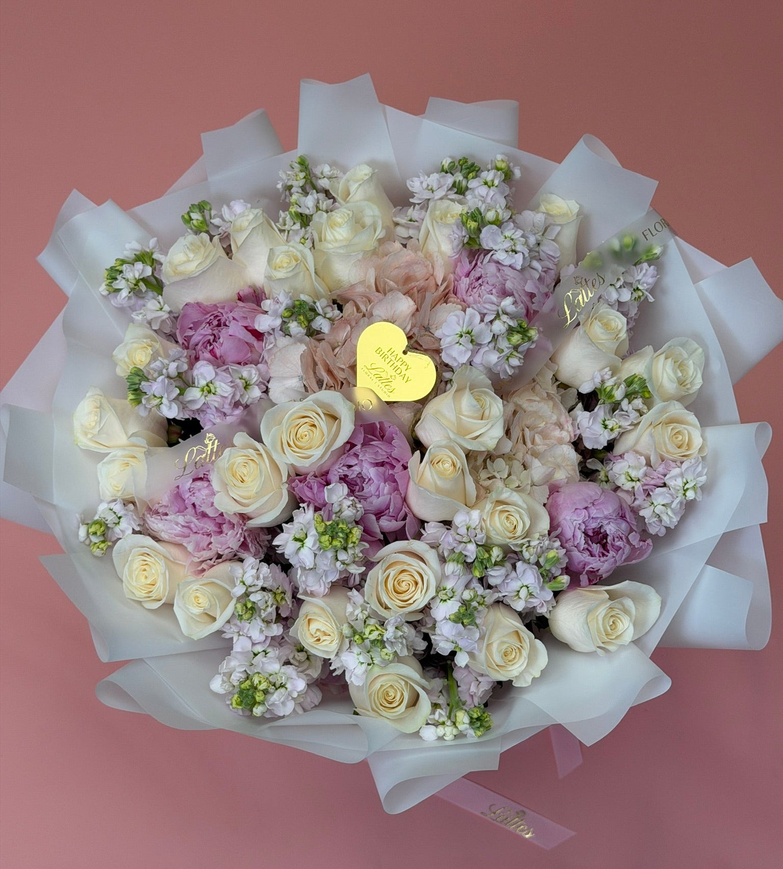 A top-down view of a large floral bouquet wrapped in white tissue paper, featuring cream and light pink roses, lavender carnations, and green filler flowers, with a small gold heart-shaped card in the center, set against a solid dusty pink background.