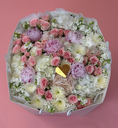 A large, round floral arrangement in a white gift box with white tissue paper, featuring pink and white flowers including roses, peonies, and chrysanthemums, with a small gold heart-shaped card in the center, set against a solid pink background.