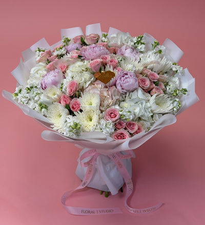 A floral bouquet wrapped in white paper and tied with a pink ribbon, featuring pink and white flowers, set against a solid pink background.