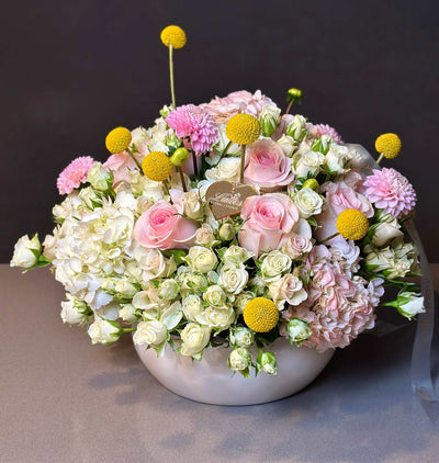 Golden Bliss Floral Arrangement with pastel pink roses, white spray roses, hydrangea, yellow craspedia, and chrysanthemums in a white vase