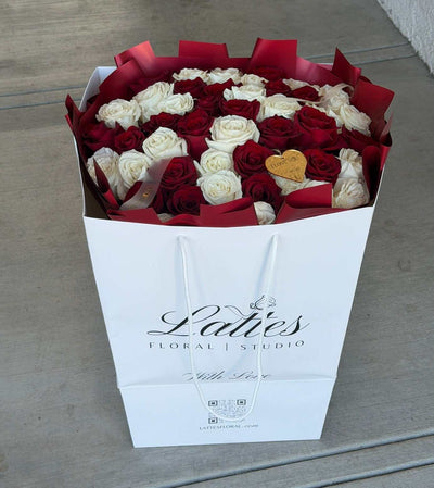 A large bouquet of red and white roses in a white gift box with red tissue paper, placed on a concrete floor. The box is branded 'Ladies Floral Studio'.