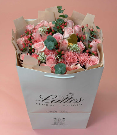 A large bouquet of pink roses, carnations, and green eucalyptus stems arranged inside a white gift bag with the brand name 'Lattes Floral Studio' printed on the front, set against a solid pink background.