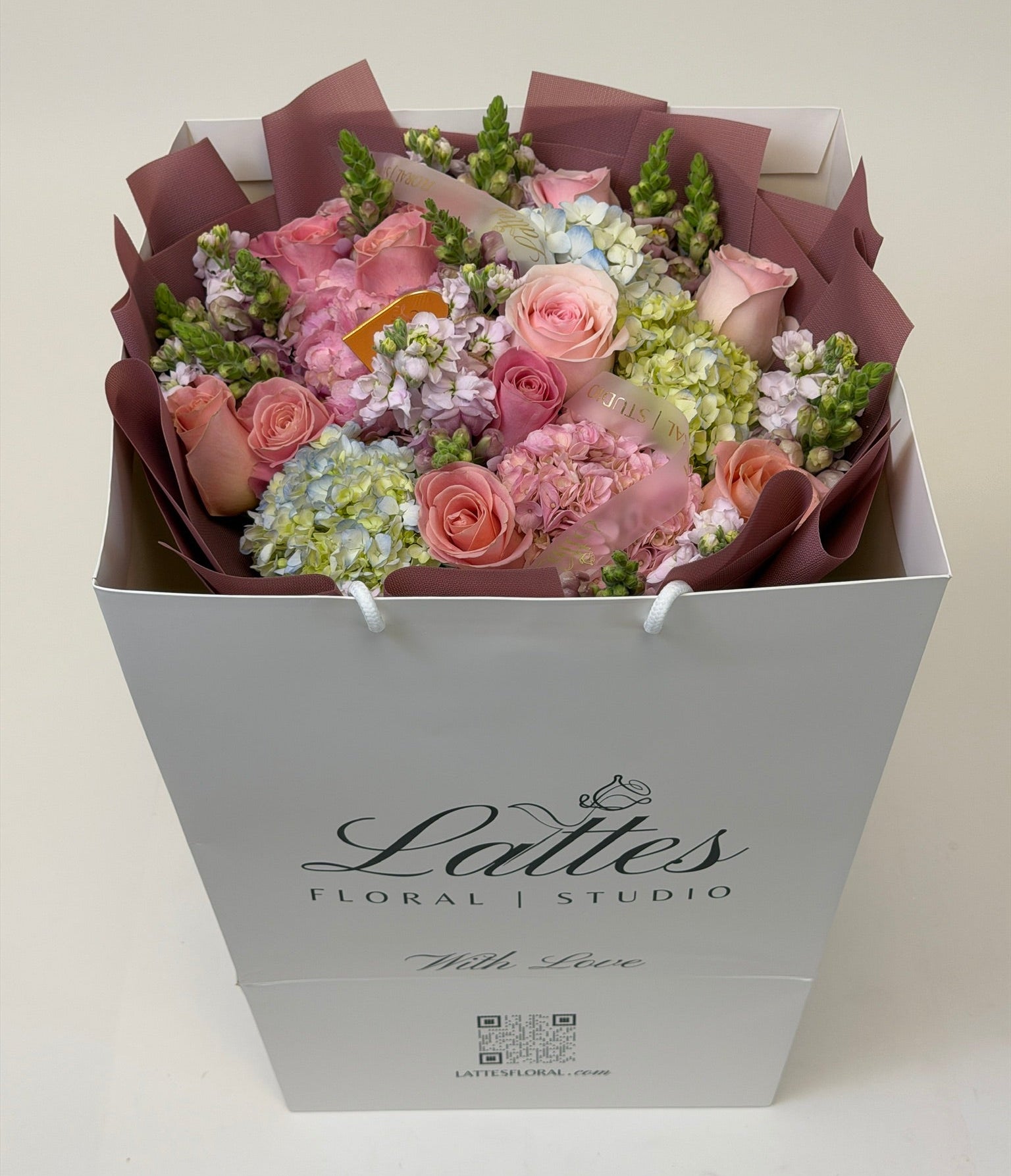 A floral bouquet in a white gift box from Lattes Floral Studio, featuring pink roses, light pink hydrangeas, green foliage, and pale blue flowers wrapped in mauve paper.