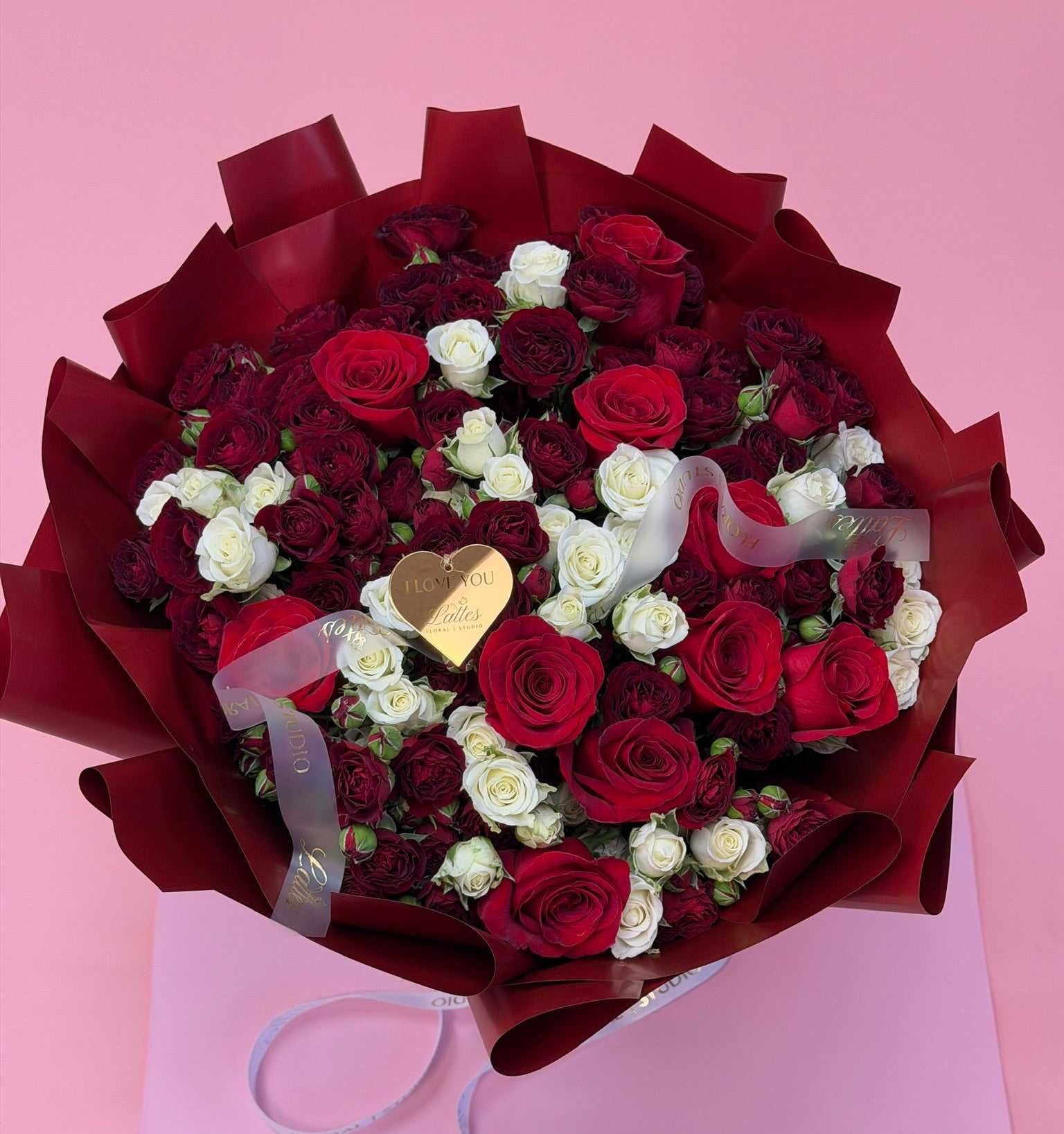 A large bouquet of red and white roses wrapped in dark red paper, with a small gold heart-shaped card in the center, set against a solid pink background.
