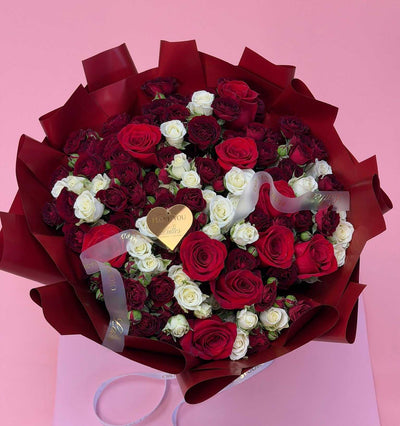 A large bouquet of red and white roses wrapped in dark red paper, with a small gold heart-shaped card in the center, set against a solid pink background.