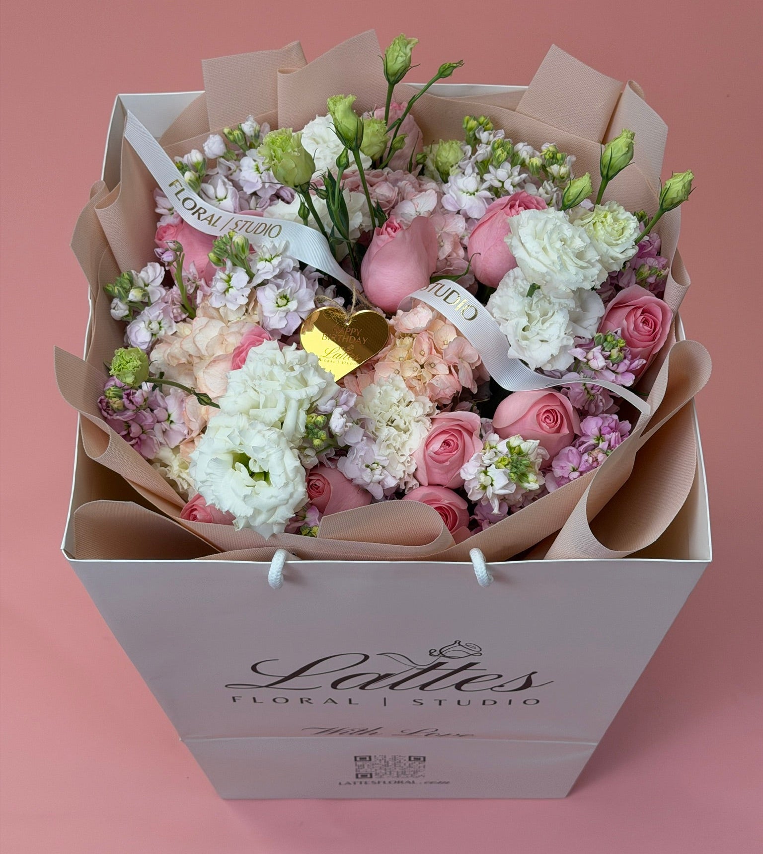 A floral bouquet in a white gift box from Lattes Floral Studio, featuring pink and white flowers, green stems, and a small gold heart accent, set against a solid pink background.