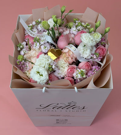A floral bouquet in a white gift box from Lattes Floral Studio, featuring pink and white flowers, green stems, and a small gold heart accent, set against a solid pink background.