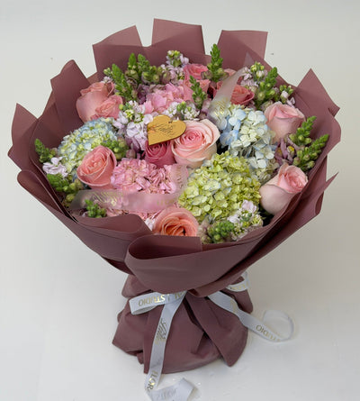 A floral bouquet wrapped in dusty mauve paper, featuring pink roses, light pink hydrangeas, green filler flowers, and a small gold heart-shaped tag, tied with a white ribbon on a plain light background.