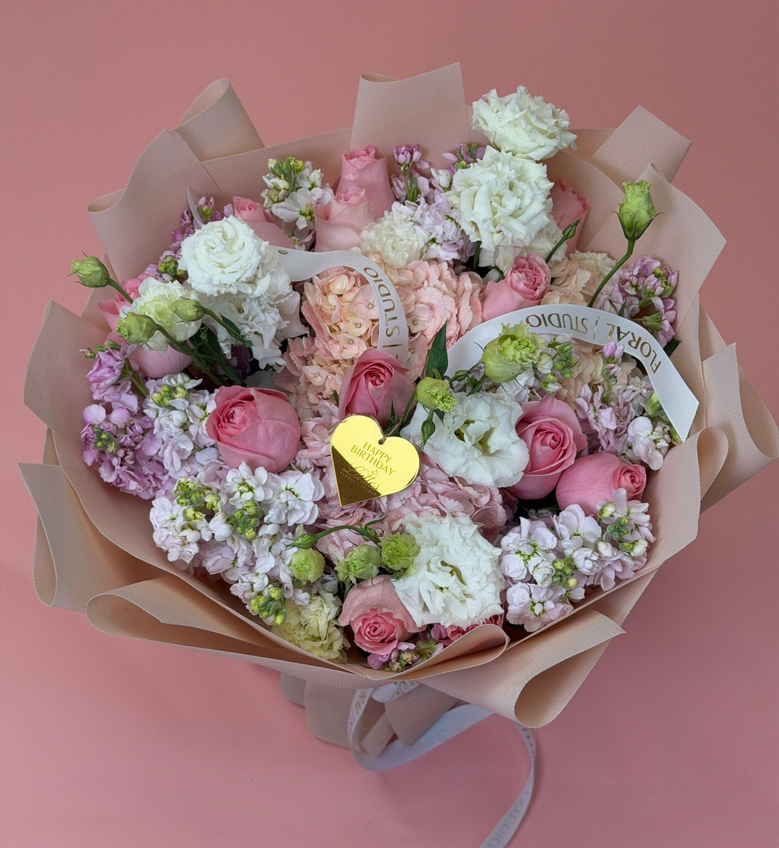 A bouquet of pink and white flowers wrapped in beige paper, featuring roses, carnations, and small purple blossoms, with a small gold heart-shaped card in the center, set against a solid pink background.