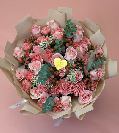 A round floral bouquet wrapped in beige paper, featuring pink roses, pink carnations, green eucalyptus leaves, and small white filler flowers, with a small yellow heart-shaped card in the center, set against a solid dusty pink background.