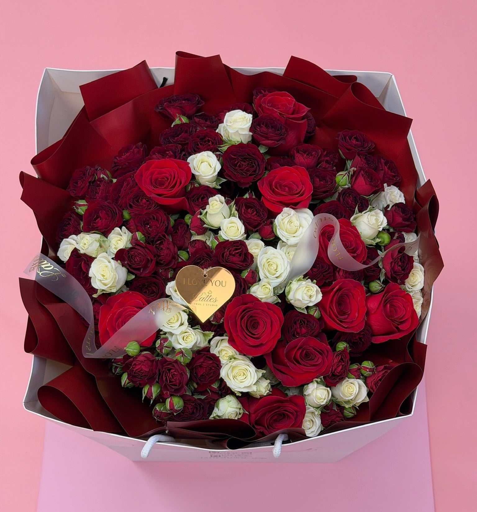 A large gift box of roses filled with deep red and white roses, wrapped in dark red paper, with a small gold heart-shaped tag, set against a solid pink background.