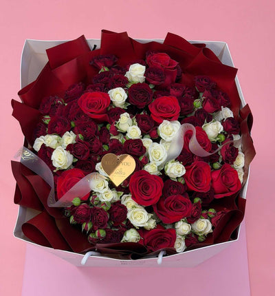 A large gift box of roses filled with deep red and white roses, wrapped in dark red paper, with a small gold heart-shaped tag, set against a solid pink background.
