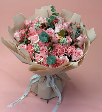 A bouquet of pink roses, carnations, and green eucalyptus stems wrapped in beige paper with a white ribbon, set against a solid pink background.