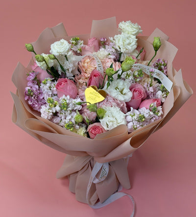A hand-tied bouquet of pink and white flowers wrapped in beige paper, featuring roses, carnations, and lilac sprigs with a small gold heart-shaped tag, set against a solid pink background.