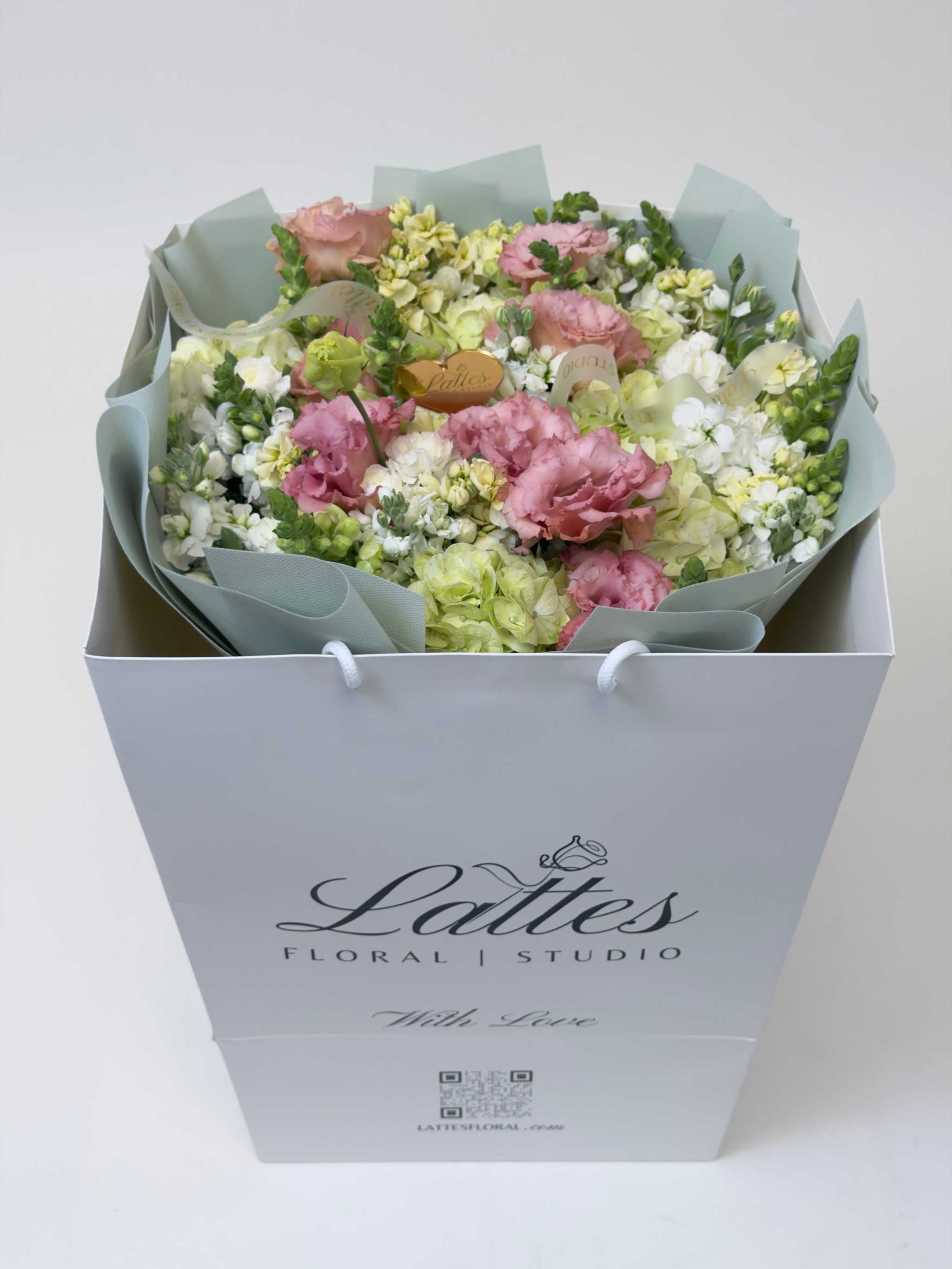 A large floral bouquet in a white gift box with the brand name 'Lalies Floral Studio' printed on the front, wrapped in pale sage green tissue paper. The bouquet features pink roses, white carnations, and light green hydrangeas with small yellow filler flowers.