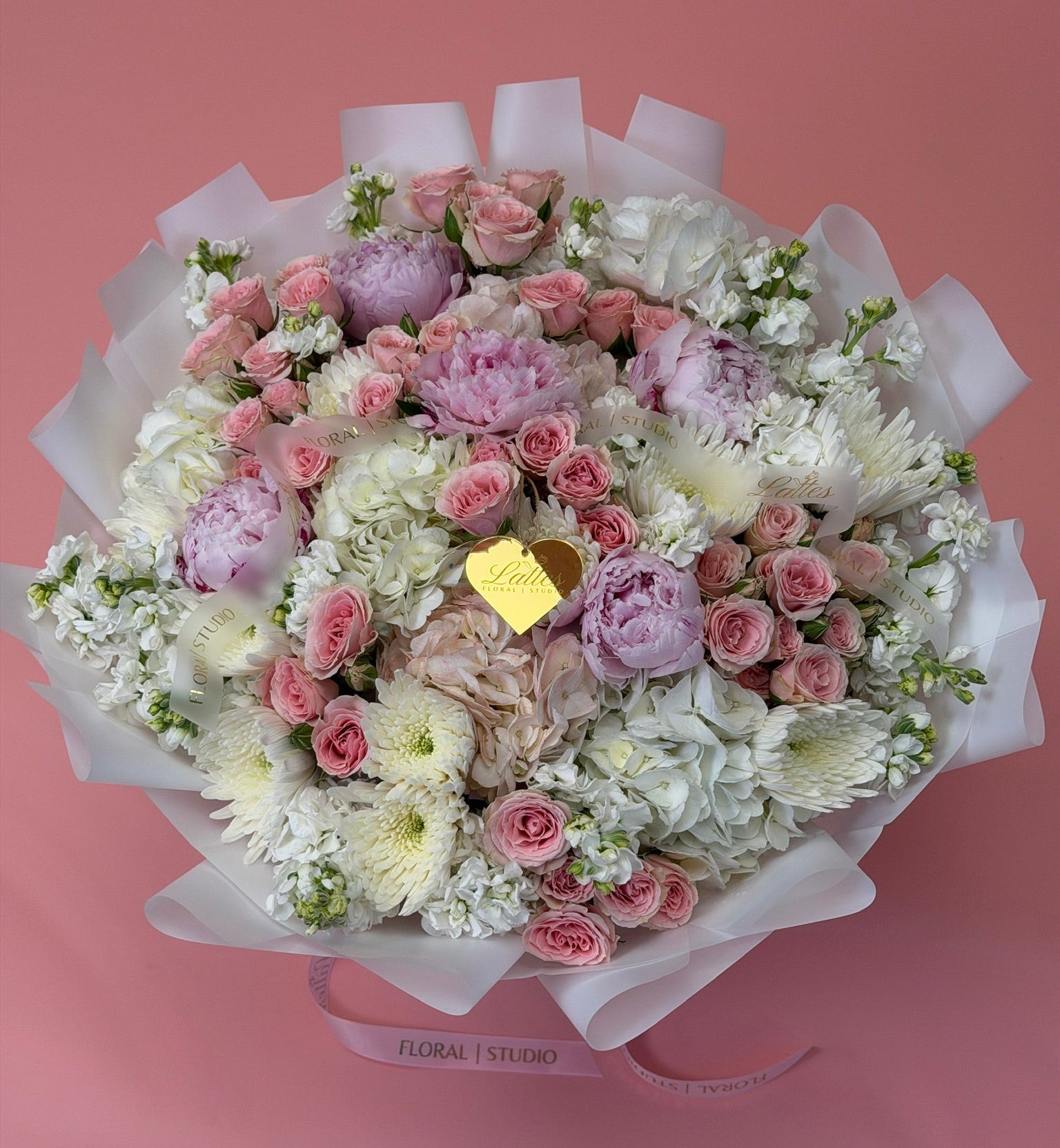 A round floral bouquet wrapped in white tissue paper, featuring pink and lavender roses, white chrysanthemums, and purple peonies, with a small gold heart-shaped tag, set against a solid pink background. 