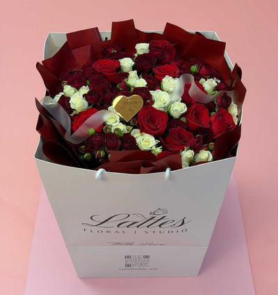 A large gift box bouquet of red and white roses wrapped in dark burgundy paper, with a small gold heart-shaped ornament in the center, presented in a white floral box printed with 'Ladies Floral Studio' on a pink background.