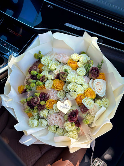 Golden Crème bouquet with yellow and white roses, lisianthus, and brown-chocolate blooms in creamy white wrapping