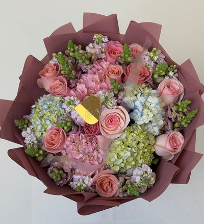 A top-down view of a floral bouquet wrapped in dark brown paper, featuring pink roses, light pink hydrangeas, pale blue hydrangeas, and green filler flowers, with a small gold heart-shaped card in the center.