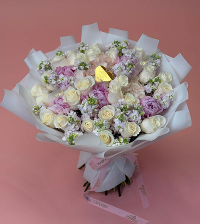 A bouquet of white and light purple roses wrapped in white tissue paper with a pink ribbon, featuring a small gold heart-shaped chocolate in the center, set against a solid pink background.