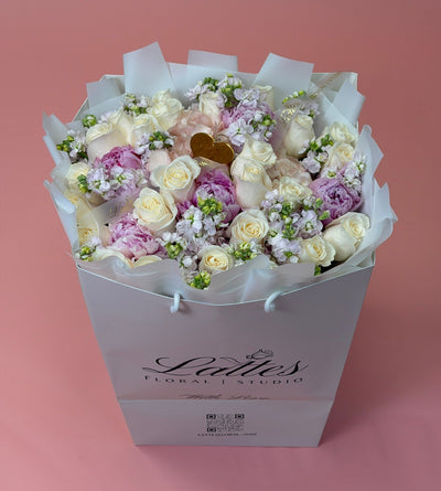 A floral bouquet in a white gift bag from Lady's Floral Studio, featuring cream and light purple roses with white filler flowers, set against a solid pink background.